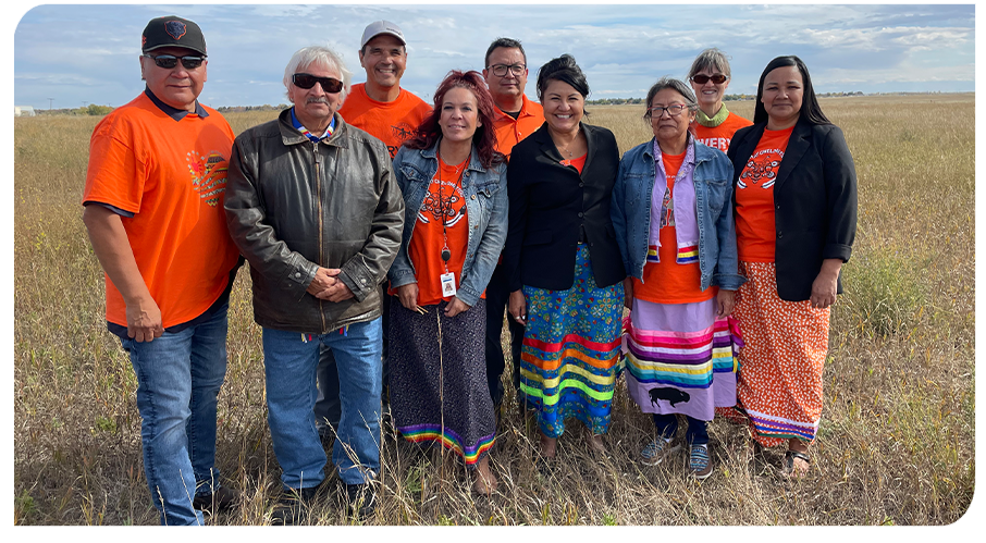 Nine people stand in a row outdoors in a golden field. Many wear orange T-shirts and/or colourful ribbon skirts.
