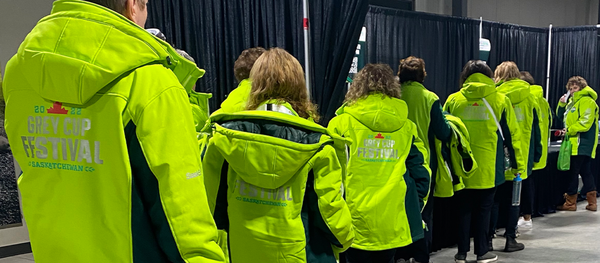 Ten volunteers all wearing matching bright green "Grey Cup Festival" jackets line up to register for their shift. 