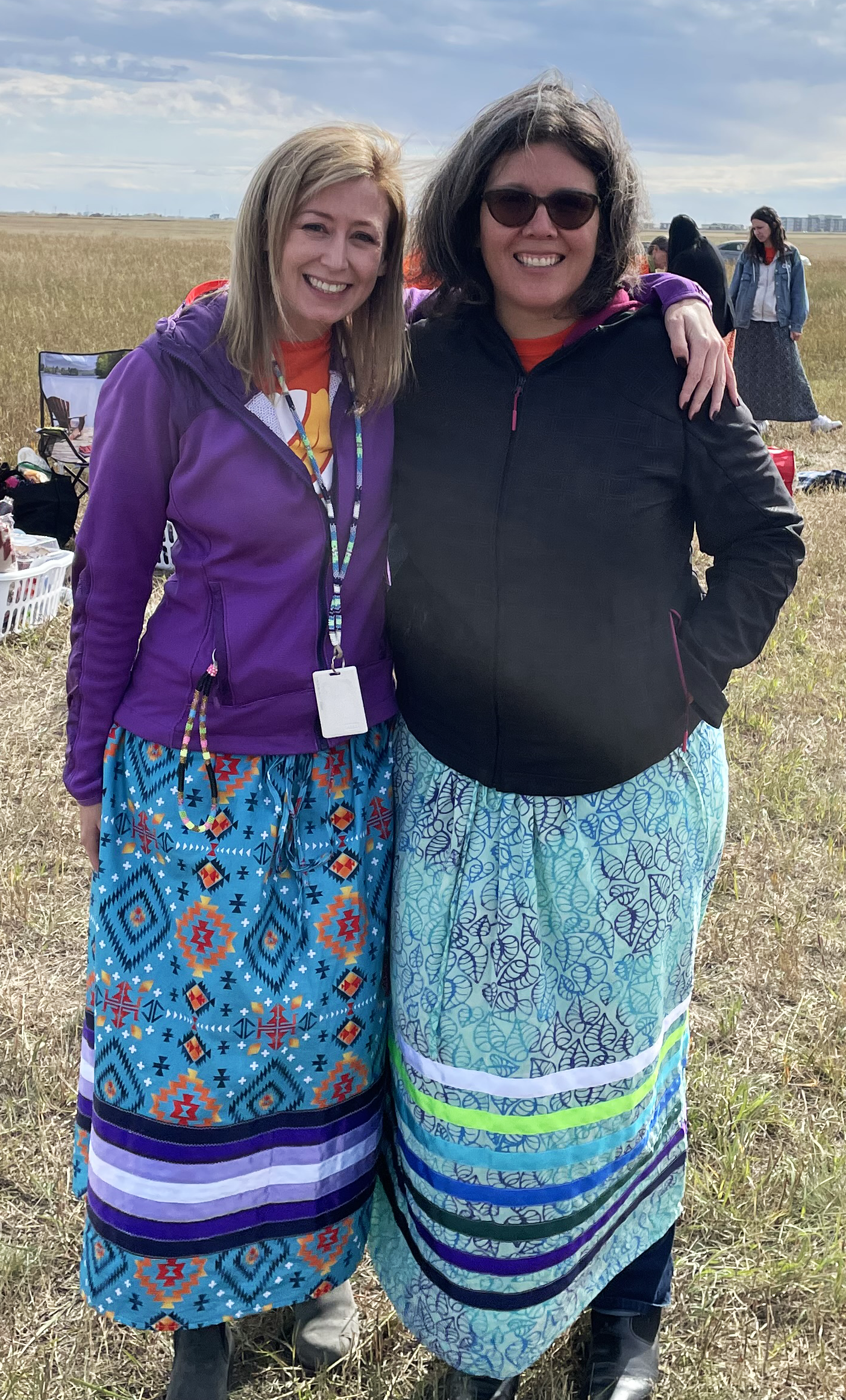 Two women stand close together smiling. Both are wearing a colourful ribbon skirt.
