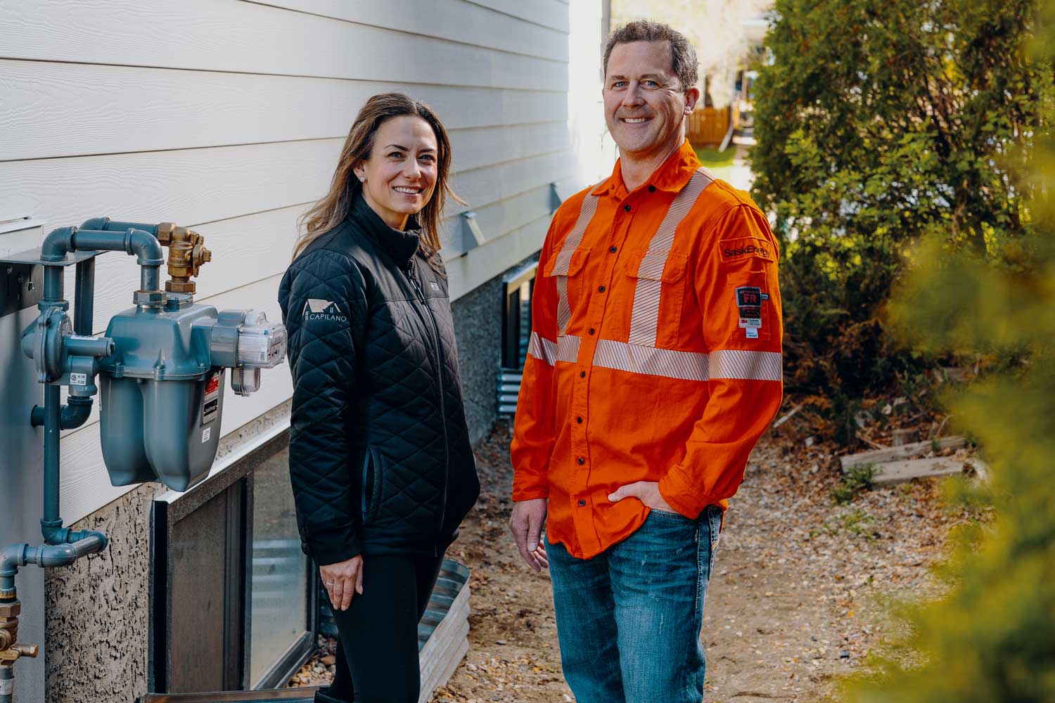 a woman with light brown hair and a black jacket, and a man in an orange safety shirt, stand next to a gas meter.