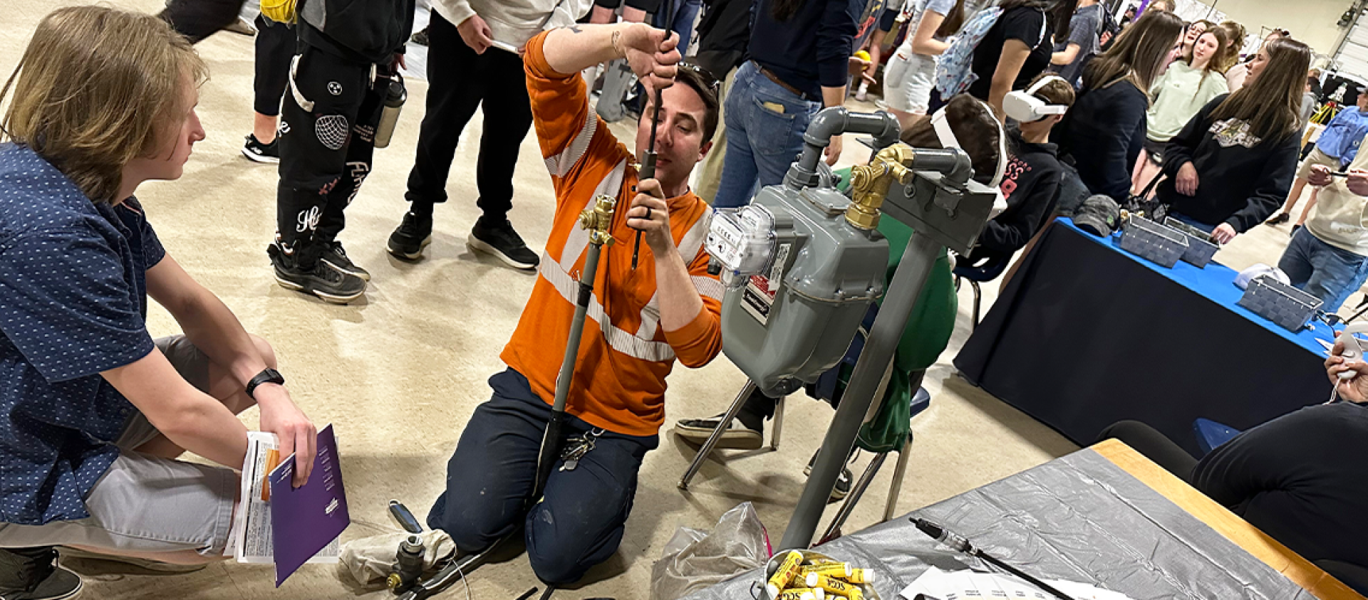 A Service Technician wearing an orange work shirt kneels next to a gas meter to do work while a student watches. 