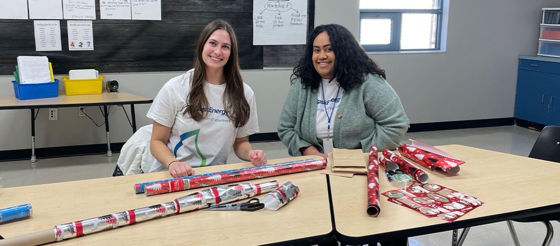 Two women with long hair smile, seated at a table with tubes of red wrapping paper in front of them.paper 