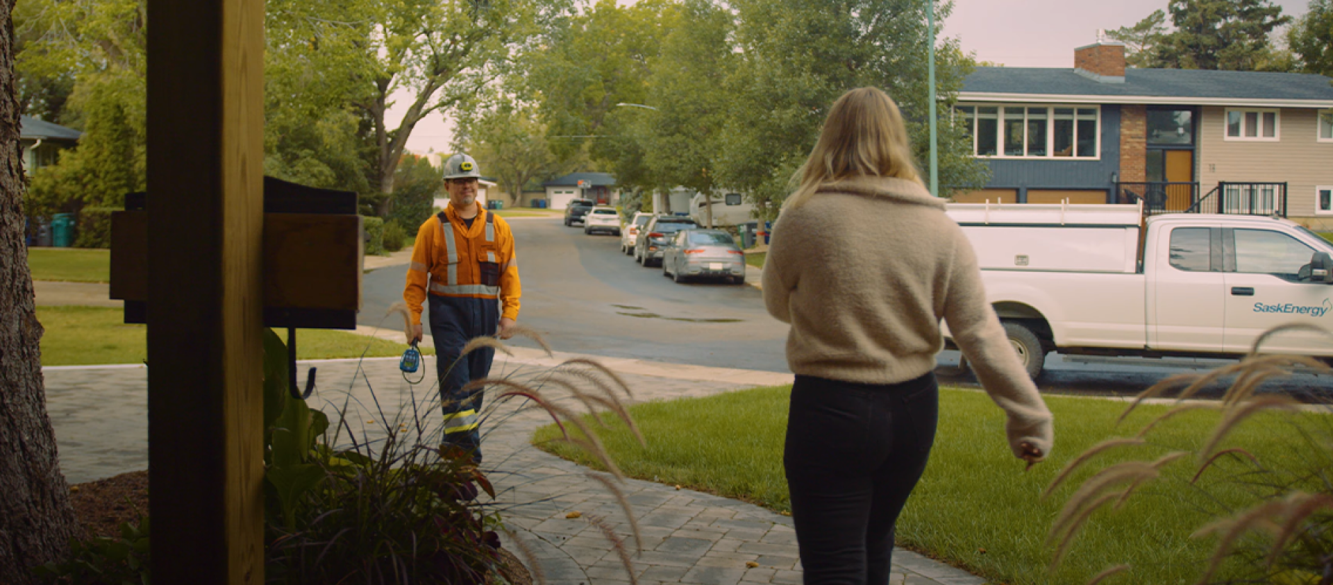 A man in a hardhat and orange work clothes walks up a pathway toward a blonde woman in a beige shirt.