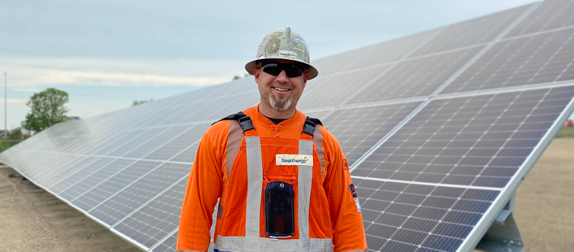 A man in a grey hardhat and orange long-sleeved safety shirt stands in front of solar panels, with blue sky and a tree beyond.