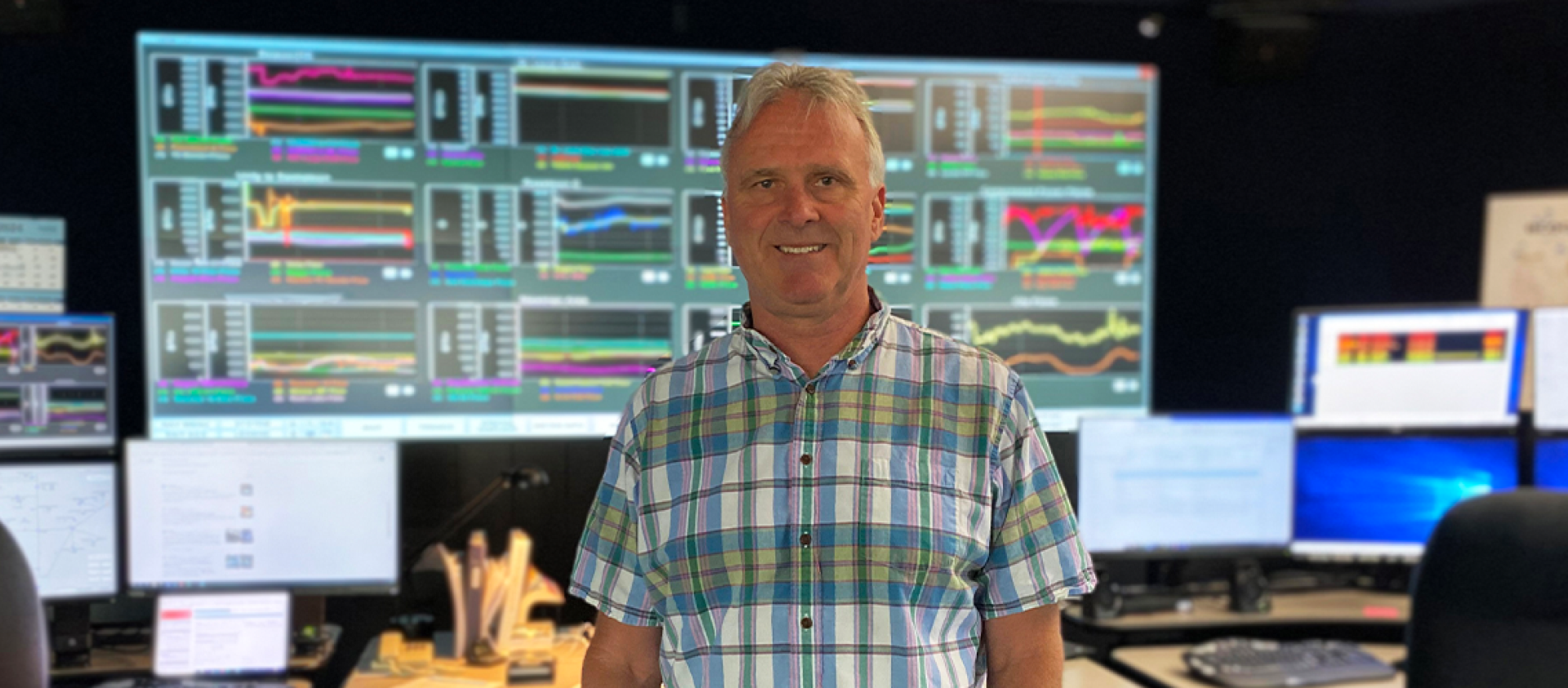 A man in a plaid shirt stands in front of colourful computer screens.