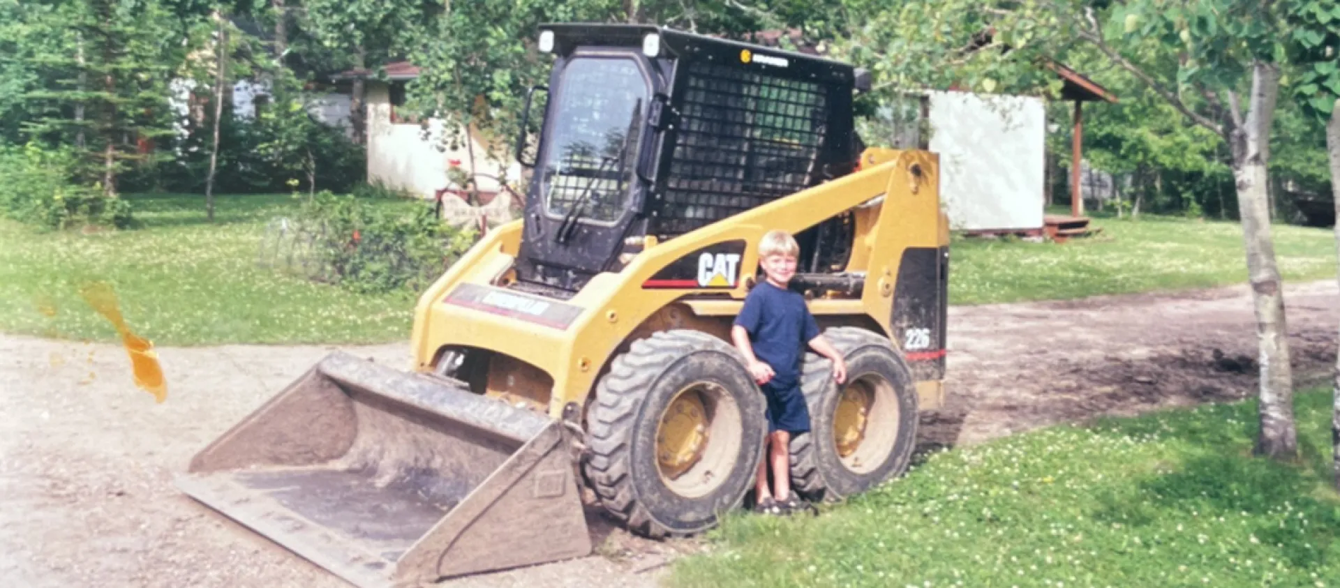 A blond child in a blue shirt stands between the large tires of a yellow construction equipment skid steer.