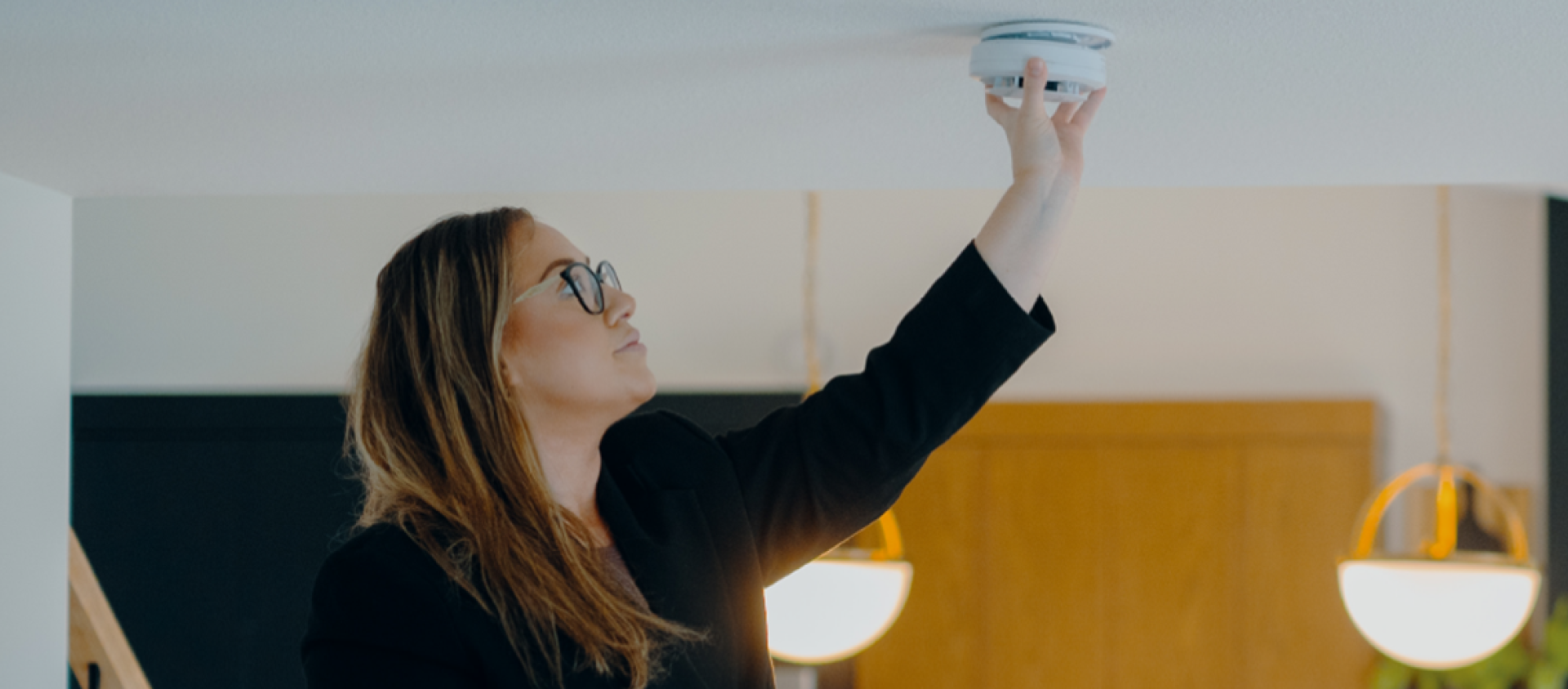 A woman with long brown hair and glasses reaches up to the ceiling to install a round CO alarm.