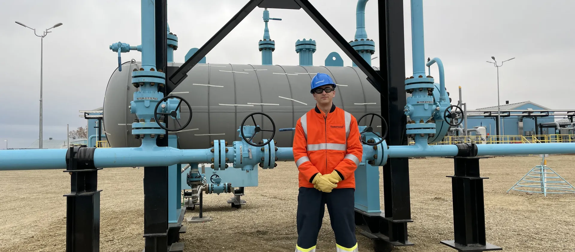 A SaskEnergy employee wearing orange high-vis safety clothing stands in front of an above-ground pipeline structure.