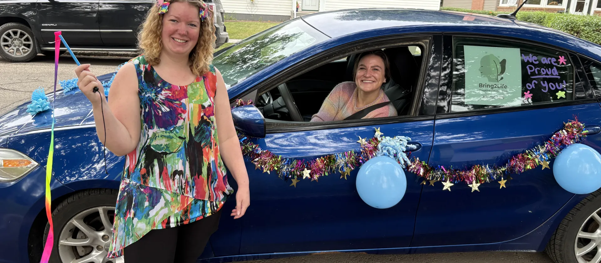 A woman stands beside a dark blue car, which is decorated in balloons and tinsel, another women sits inside the car.