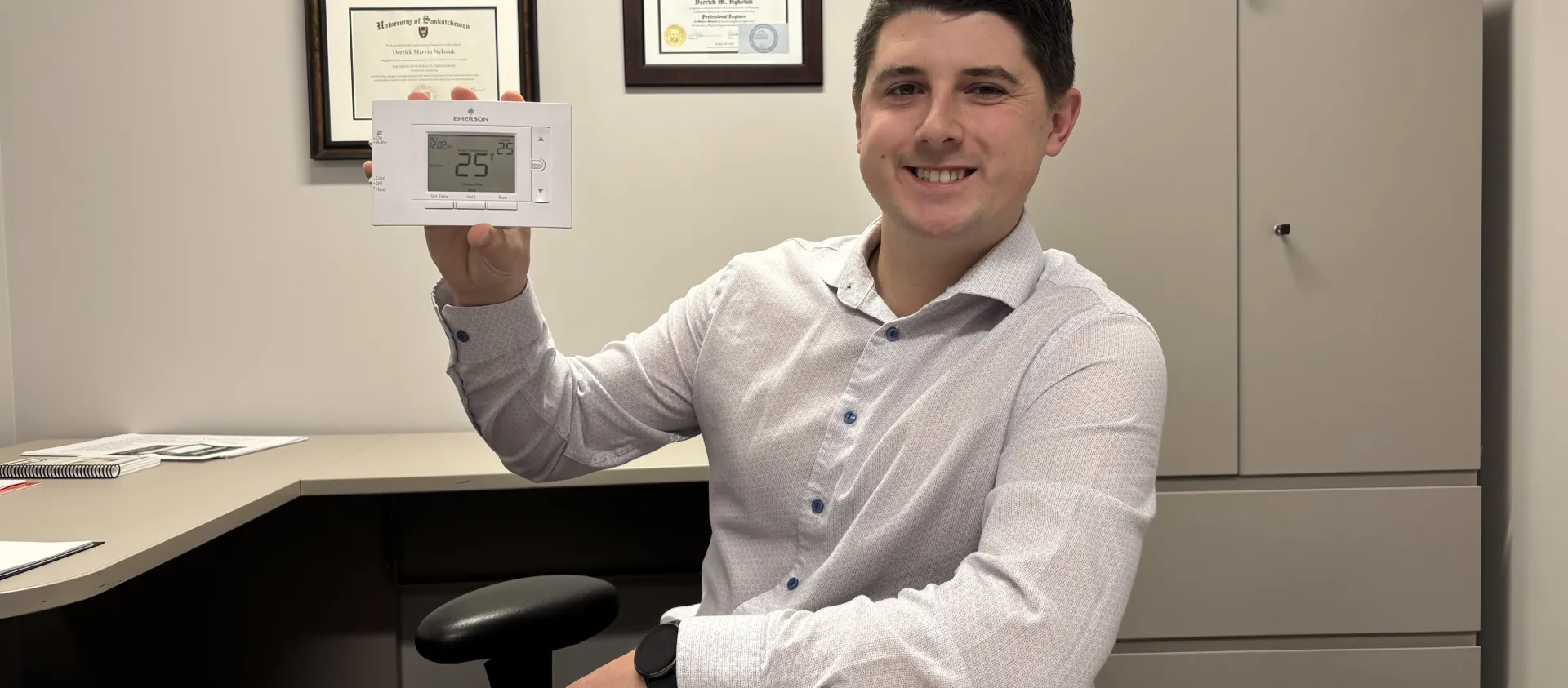 A man sits at a desk holding up a programmable thermostat.