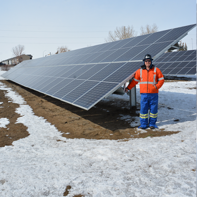 A SaskEnergy employee wearing an orange coat, blue pants and black hardhat, stands in front of a solar panel installation. Some snow is on the ground, with brown grass peeking through.