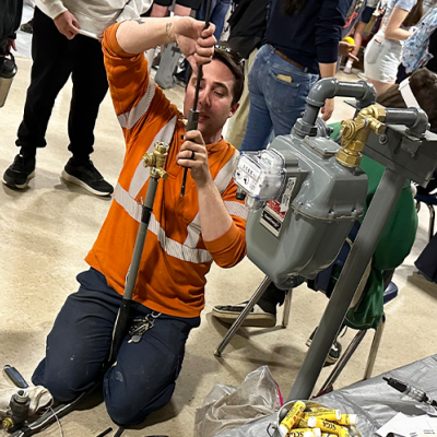 A Service Technician wearing an orange work shirt kneels next to a gas meter to do work while a student watches. 
