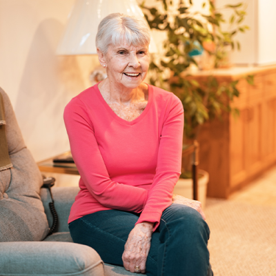 A woman with short light grey hair sits on a chair, arms loosely crossed. She's wearing jeans and a dark pink long-sleeved top.