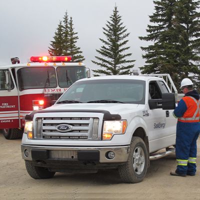 A man wearing orange and blue coveralls stands near a SaskEnergy branded white truck, with a red fire engine parked behind.