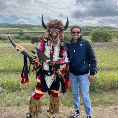 A person in traditional Indigenous dress poses outside with a person in blue jeans and a jacket. 