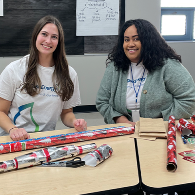 Two women with long hair smile, seated at a table with tubes of red wrapping paper in front of them.paper 