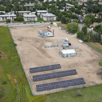 aerial image - three rows of solar panels at centre, surrounded by light brown earth, grass and trees, houses and roads. 