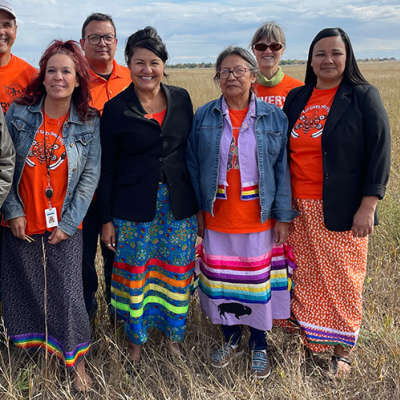 Six people stand in a row in a golden field. Four women in front wear colourful skirts and many wear orange T-shirts.