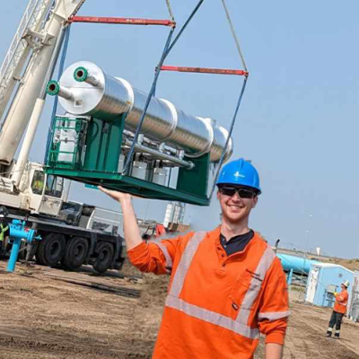 A man wearing an orange workshirt and blue hard hat holds up his arm, to make it appear he is lifting a large silver line heater on a crane in the background.