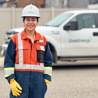 A woman in an orange and blue work uniform, white hard hat and yellow gloves stands in front of a SaskEnergy truck.