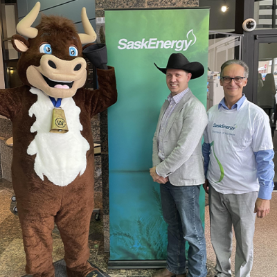 A brown and white cow mascot stands next to a man in a black cowboy hat and another man wearing glasses. A green and blue banner with the SaskEnergy logo is behind them. 