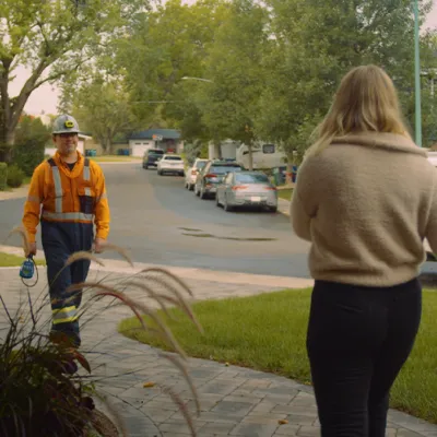 A man in a hardhat and orange work clothes walks up a pathway toward a blonde woman in a beige shirt.