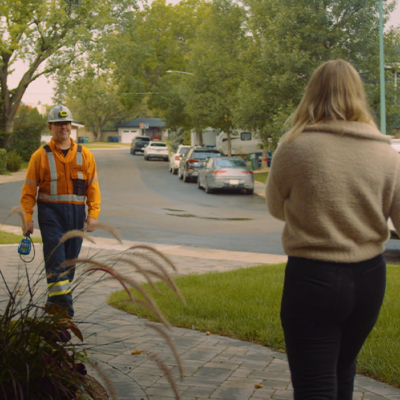 A man in a hardhat and orange work clothes walks up a pathway toward a blonde woman in a beige shirt.