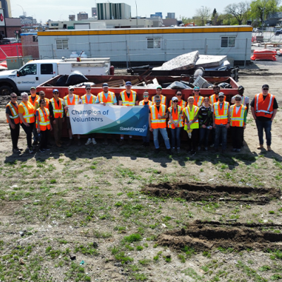 A group of about 20 people in orange safety vests stand in front of a large red dumpster. A few people hold a large banner that says "Champion of Volunteers."
