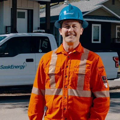 A man wearing a blue hard hat and orange safety jacket stands smiling in front of a white SaskEnergy pickup truck and two houses.