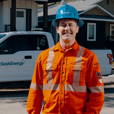 A man wearing a blue hard hat and orange safety jacket stands smiling in front of a white SaskEnergy pickup truck and two houses.