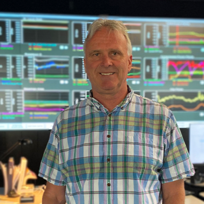 A man in a plaid shirt stands in front of colourful computer screens.