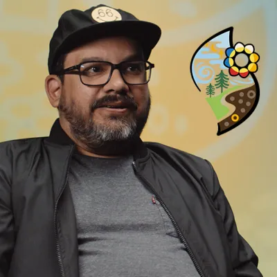 A man with a short beard and glasses in a grey T-shirt, black jacket and ballcap. A drawing of a flower, trees earth and sky scene floats on the right.