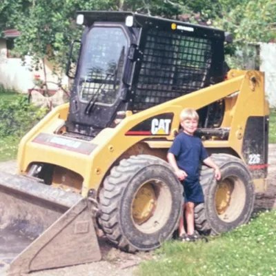 A blond child in a blue shirt stands between the large tires of a yellow construction equipment skid steer.
