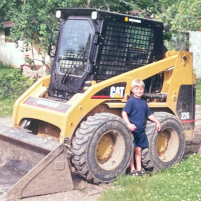 A blond child in a blue shirt stands between the large tires of a yellow construction equipment skid steer.