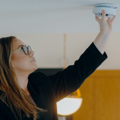 A woman with long brown hair and glasses reaches up to the ceiling to install a round CO alarm.