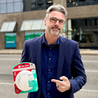 A man with short grey hair, glasses and beard in a blue suit holds a CO alarm