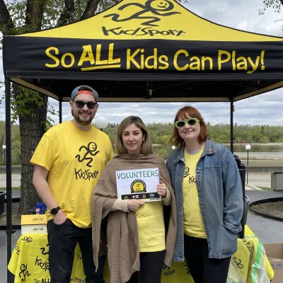 Three people stand in front of a KidSport tent