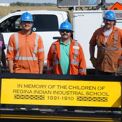 Three men in orange work shirts and blue hard hats stand behind a yellow bench.
