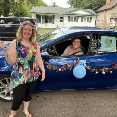 A woman stands beside a dark blue car, which is decorated in balloons and tinsel, another women sits inside the car.
