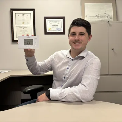 A man sits at a desk holding up a programmable thermostat.