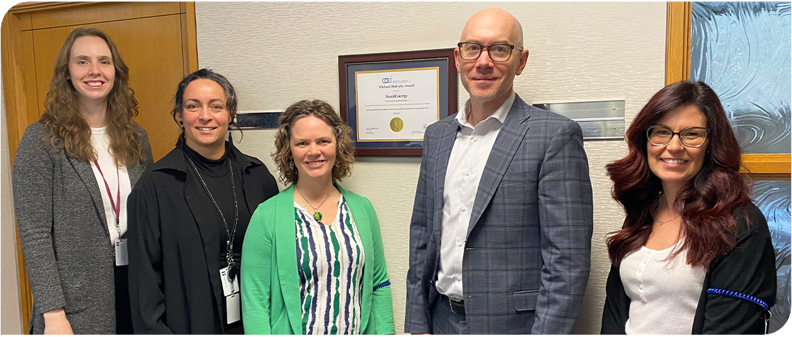 Five SaskEnergy employees stand in a row smiling, with the framed award certificate on the wall behind them.