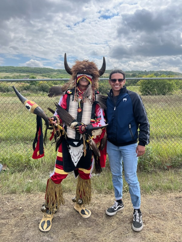 A person in traditional Indigenous dress poses outside with a person in blue jeans and a jacket. 