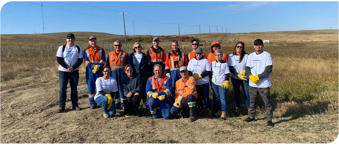 16 people - many wearing orange work clothes - stand or kneel in a posed group photo, with blue sky above and brown grass around them