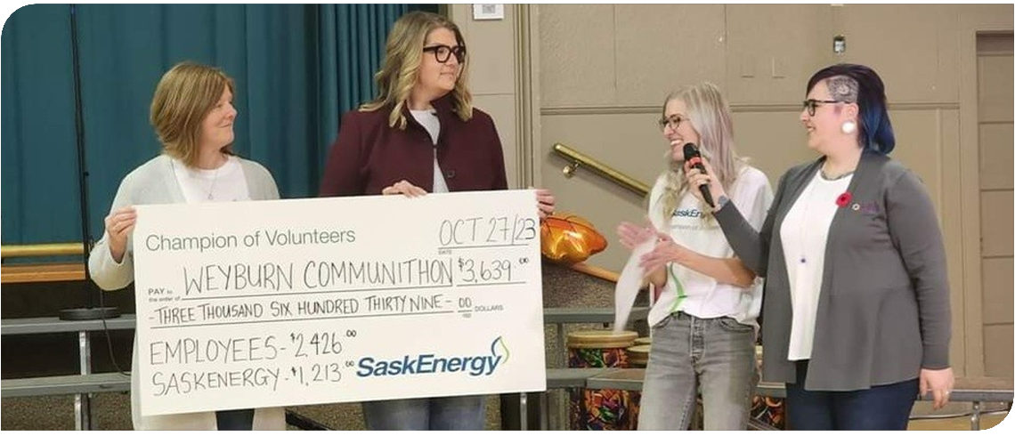 Two people hold a giant novelty cheque, while a person holds a microphone to another person's mouth.