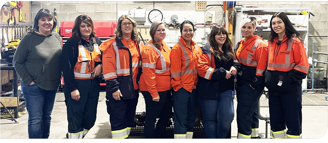 Eight women stand in a row in a shop with tools behind. Seven of them are wearing orange and navy work clothes. 