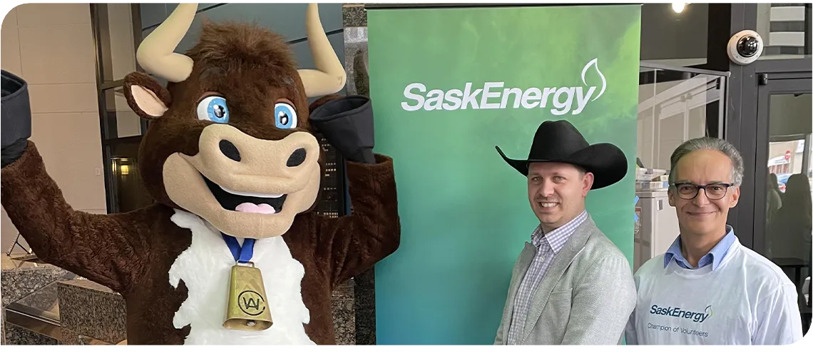 A brown and white cow mascot stands next to a man in a black cowboy hat and another man wearing glasses. A green and blue banner with the SaskEnergy logo is behind them.