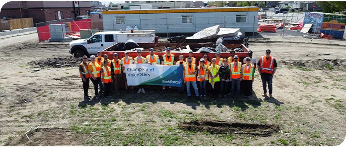 About 20 people in orange safety vests stand in front of a large red dumpster. A few people hold a banner that says "Champion of Volunteers." 