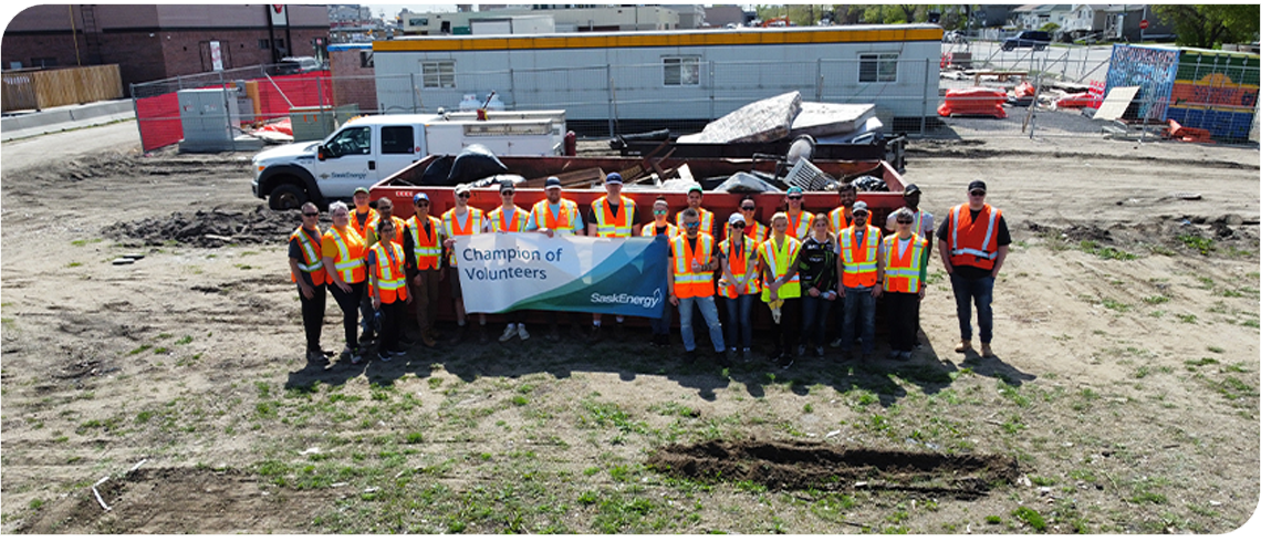 About 20 people in orange safety vests stand in front of a large red dumpster. A few people hold a banner that says "Champion of Volunteers." 