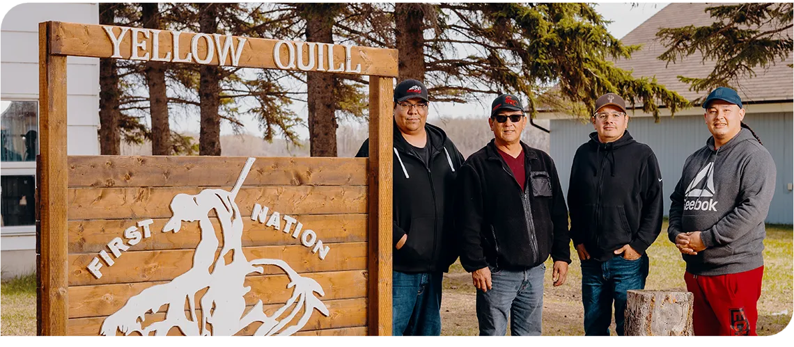 Four men stand in a row outdoors, next to a large wooden sign with the words Yellow Quill First Nation.