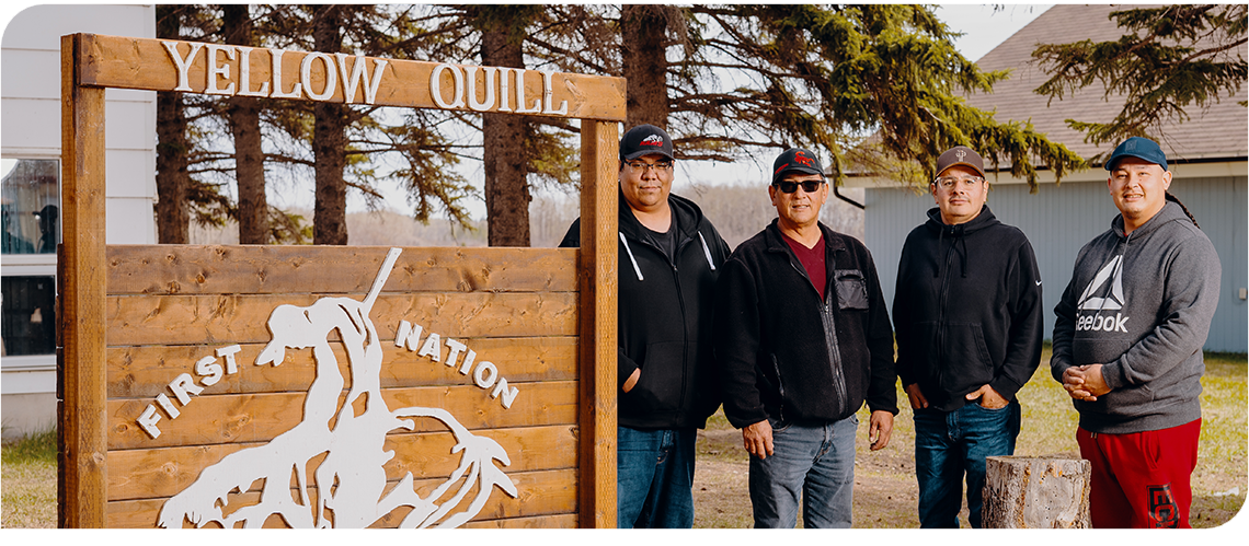 Four men stand in a row outdoors, next to a large wooden sign with the words Yellow Quill First Nation.